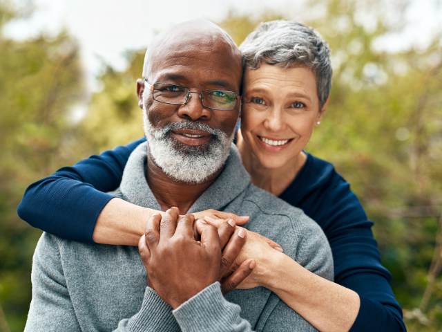 A middle-aged woman with short gray hair in a navy blue sweater wrapping her arms around a middle-aged man in a light blue sweater with a white beard and glasses